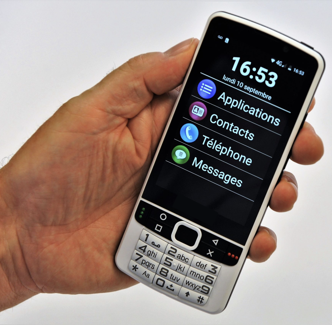 Téléphone portable en main avec, affiché à l'écran, des fonctionnalités applications, contacts, téléphone et messages. Un petit clavier à touches chiffres noirs sur fond gris