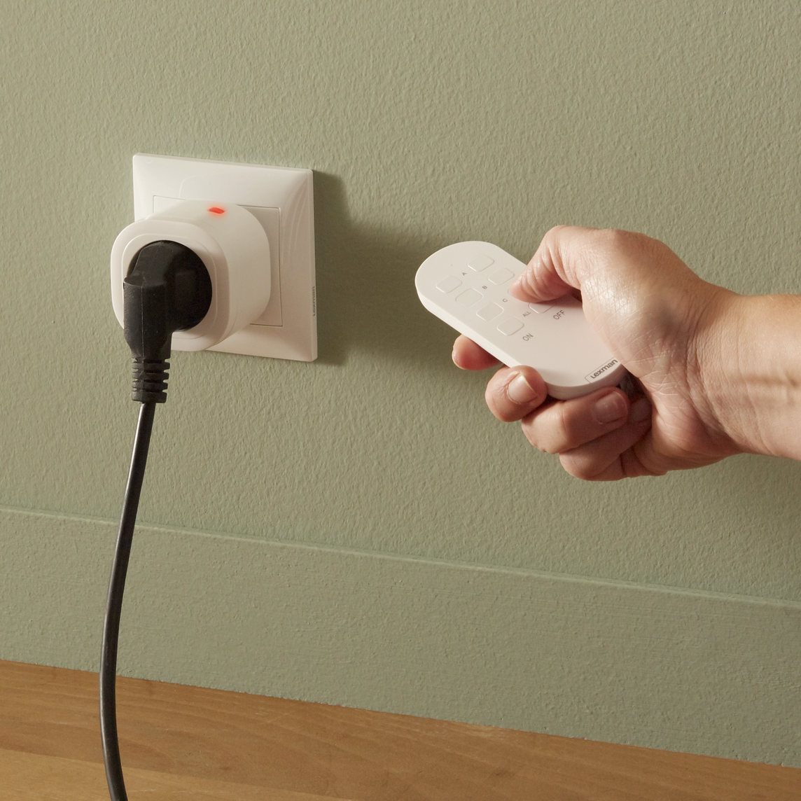 Câble noire branchée sur une prise dédiée télécommandable, elle-même branchée sur une prise murale secteur. Une main tenant une petite télécommande avec le pouce en appui sur un bouton. La télécommande est dirigée vers la câble et prise.