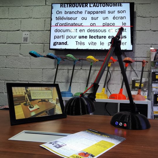 Sur la photo, un télé-agrandisseur se présente sous la forme d’une lampe-loupe noire montée sur pied, posée sur une table en bois. À côté, un écran affiche un texte agrandi, facilitant ainsi la lecture. Le dispositif fonctionne en plaçant un document sous la lampe, qui capture et diffuse l’image en version grossie sur l’écran. En arrière-plan, on aperçoit plusieurs lampes similaires de différentes couleurs, alignées contre un mur gris, suggérant un espace de démonstration ou de vente de solutions d’aide à la lecture pour personnes malvoyantes.